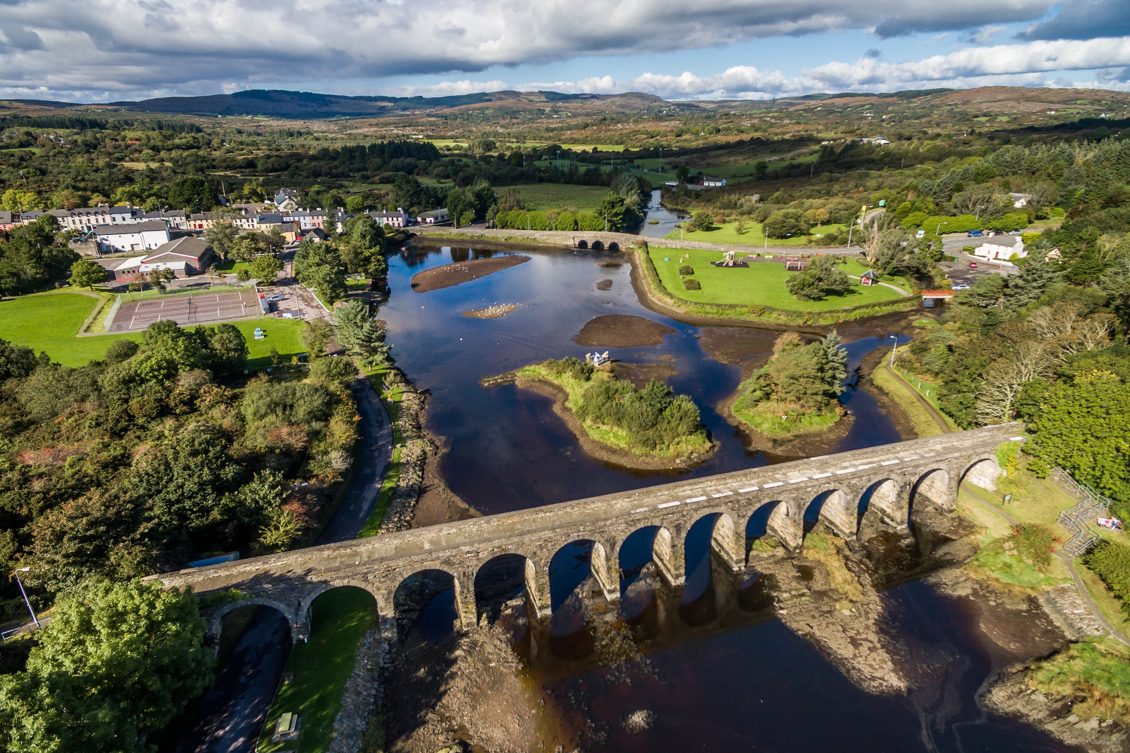 Wide aerial view across Ballydehob’s stone railway viaduct, harbour, village and the surrounding West Cork hills