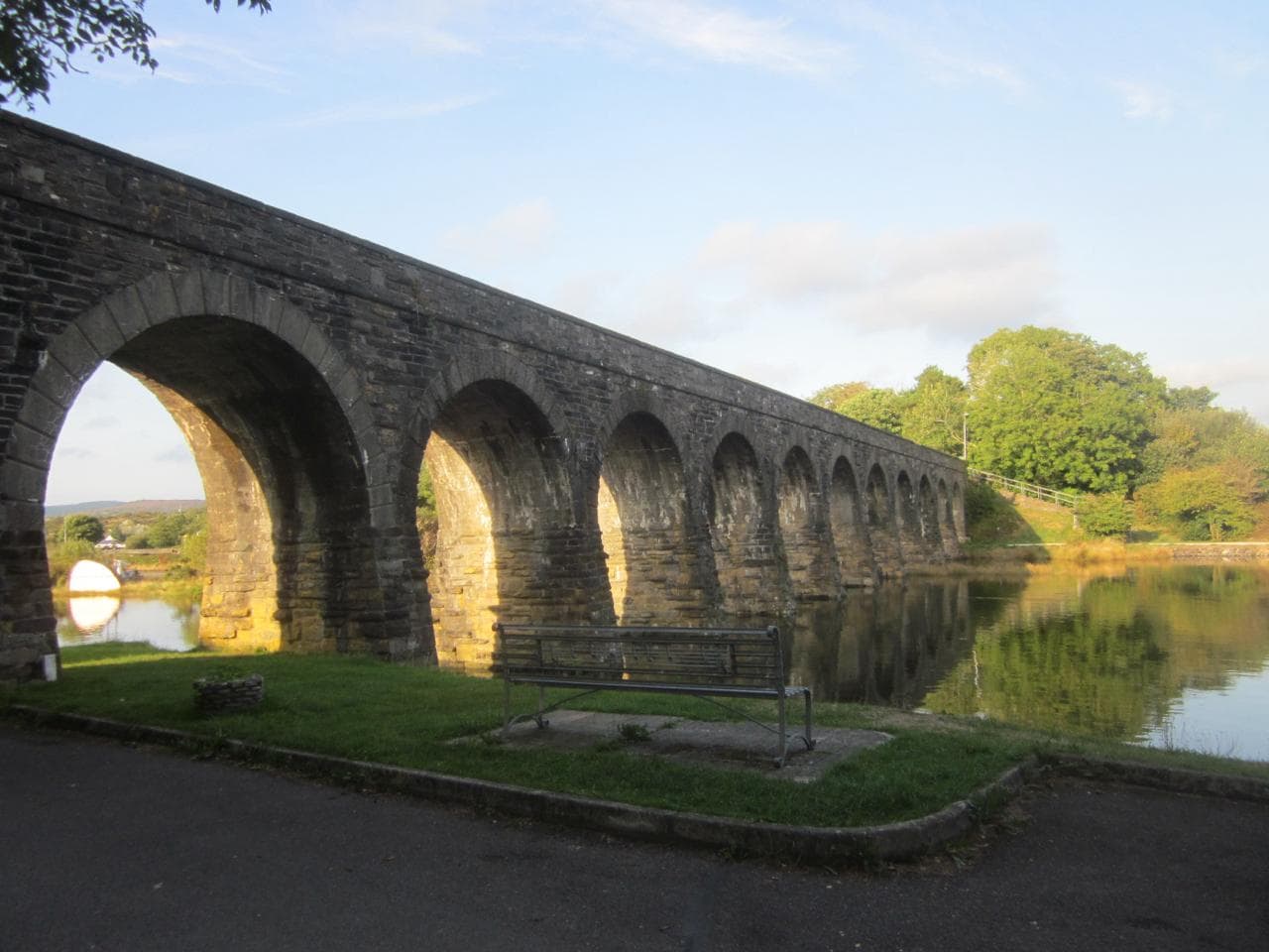 Ballydehob’s stone railway bridge in warm sunlight, with a bench beside the water